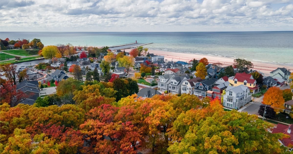 Aerial view of a coastal town with colorful autumn trees, houses, a beach, and a pier extending into the ocean under a partly cloudy sky.
