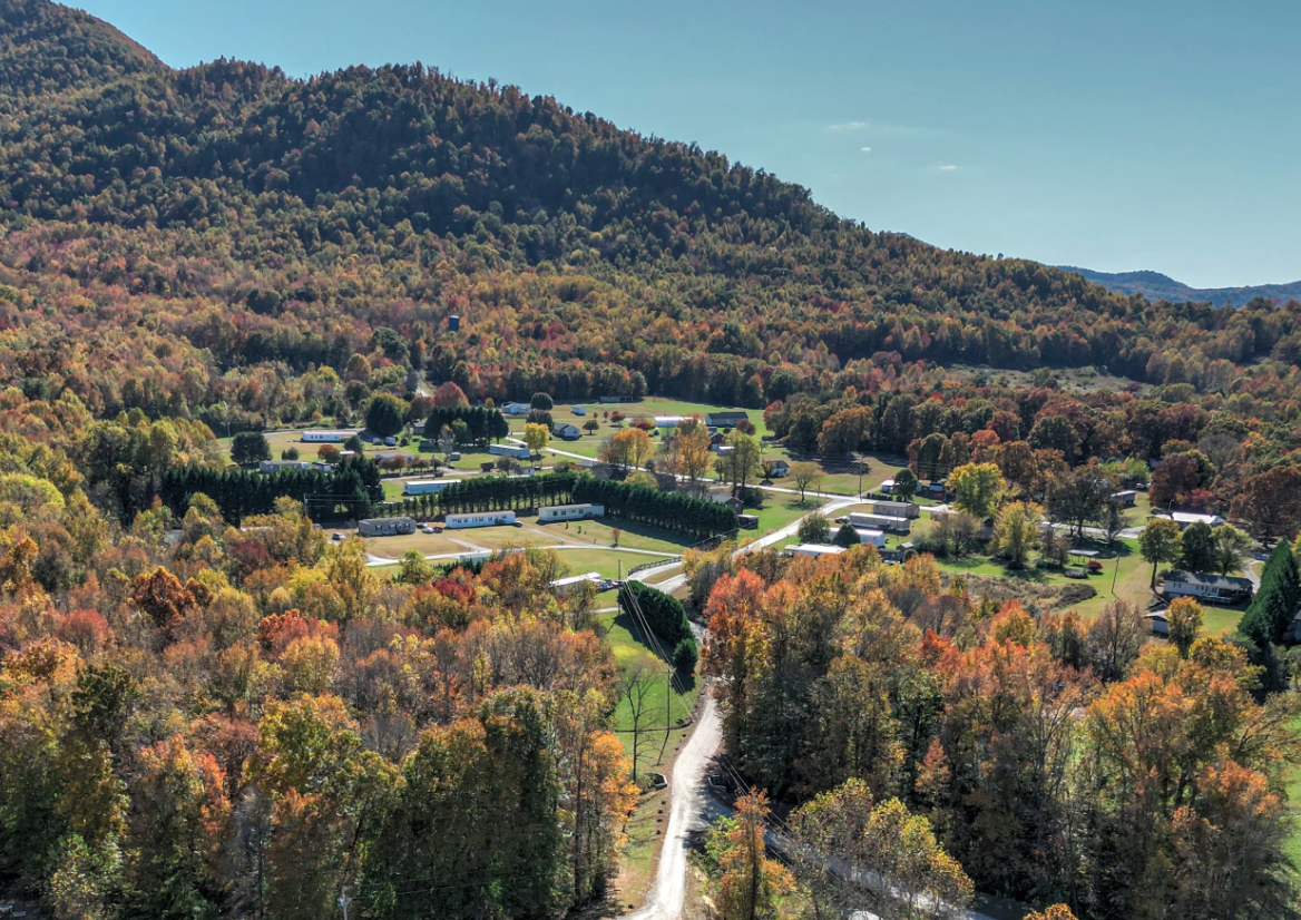 Aerial view of a rural manufactured home community surrounded by colorful fall foliage, with a mountainous background, roads, and houses.