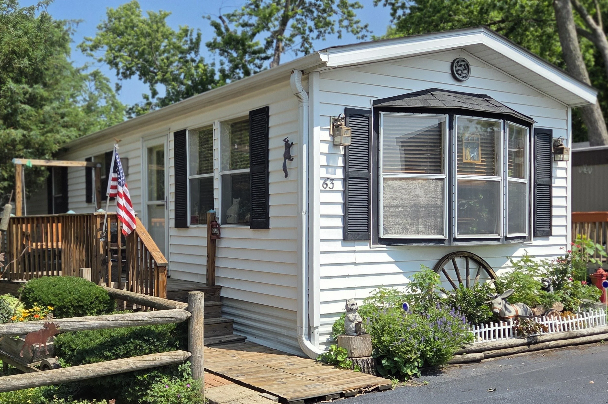 White single-story house with black shutters, a bay window, a small front porch with wooden stairs, surrounded by flowers, plants, and garden decorations, and a flag on a pole near the entrance.