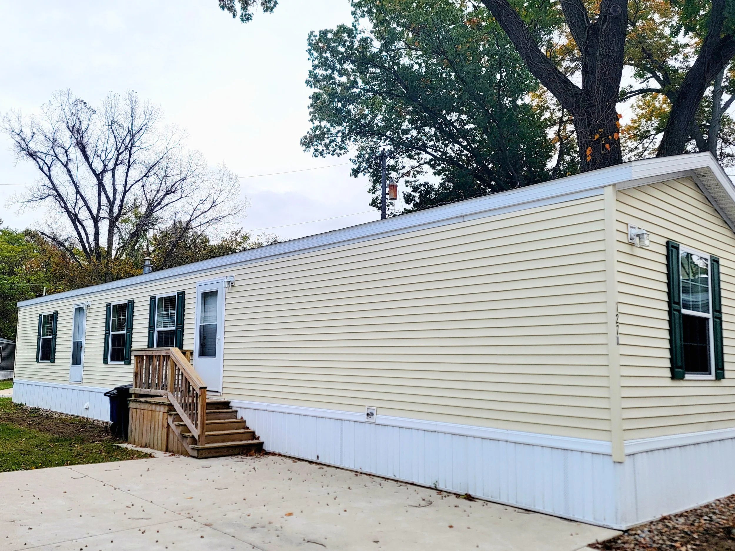 Single-story manufactured home with beige vinyl siding, green shutters, a white door and small wooden steps leading to the entrance, located on a concrete slab with a grassy yard and tall trees in the background.