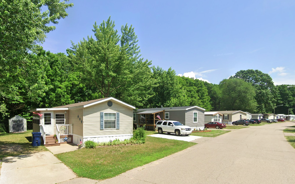 Residential mobile home community with paved road, parked cars, lush green trees, and clear blue sky.