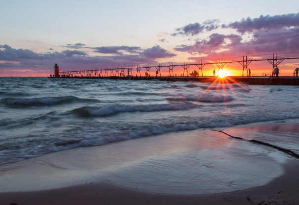 Beach with waves, a pier extending into the water, a lighthouse at the end of the pier, and a sunset in the background with colorful sky and clouds.