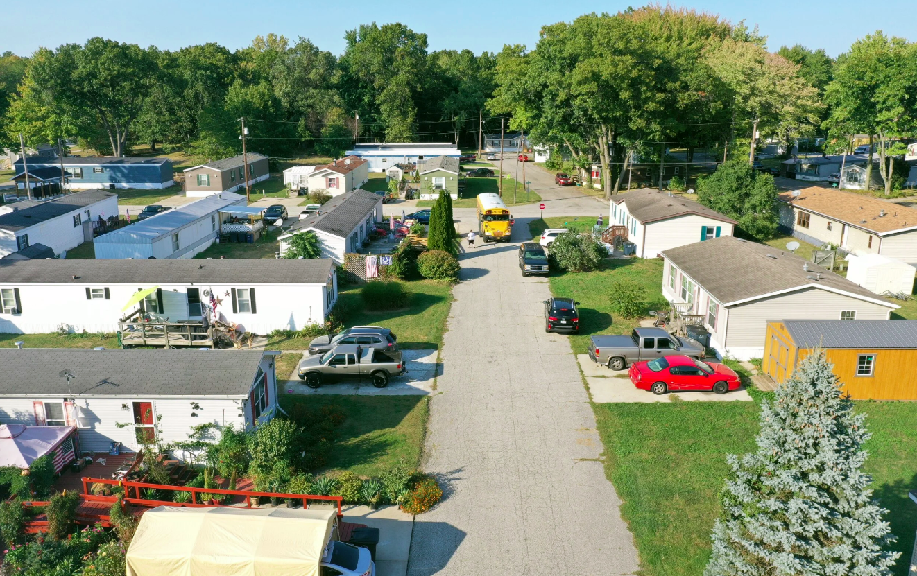 An aerial view of a residential neighborhood with multiple single-story houses, parked cars along a gravel street, trees, and a school bus at a crosswalk.