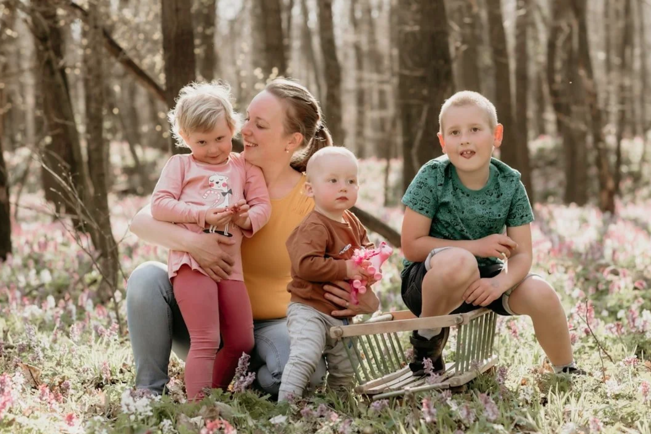 A woman with three young children in a forest with pink flowers. The woman is smiling and crouching with the children around her. One child is sitting in a small wooden wagon, and the others are standing or sitting nearby, holding toys and flowers.