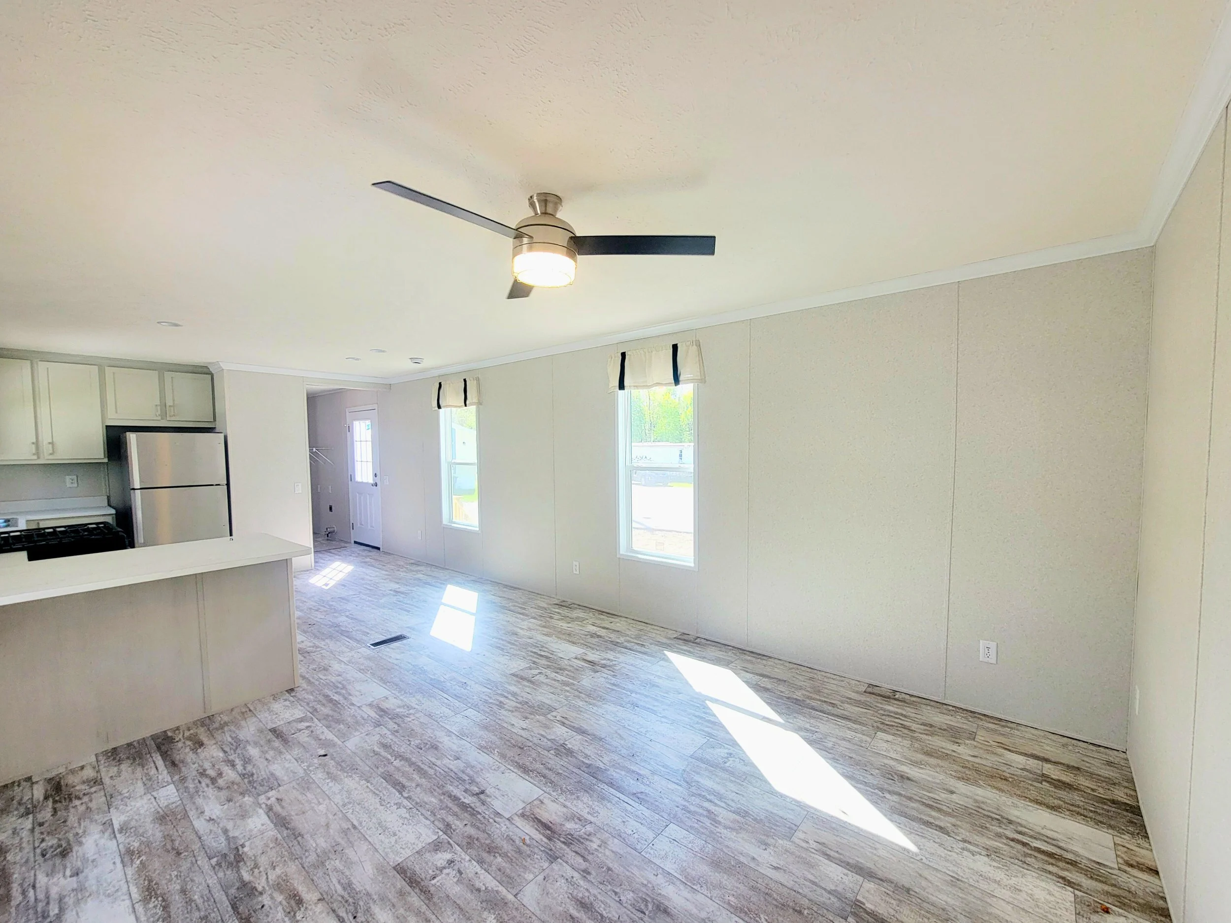 Empty living room with light-colored walls, wood-like flooring, ceiling fan with light, and windows letting in natural light.