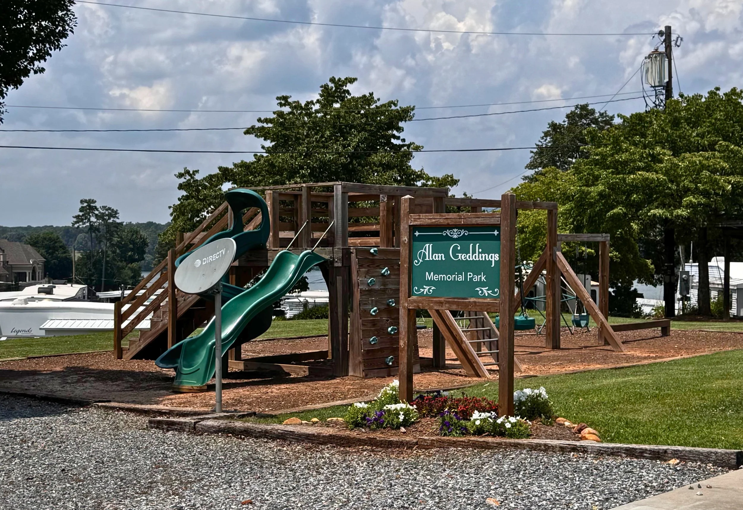 Children's playground at Alan Geddings Memorial Park, featuring a climbing structure, slide, swings, and a sign with the park's name in the foreground, with trees and blue sky in the background.