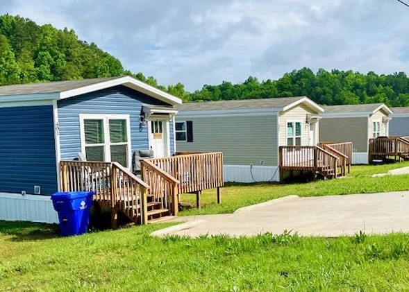Three manufactured homes with small wooden decks are arranged in a row in a residential area, with a grassy lawn and a paved driveway in front. The homes are blue, beige, and light green, with trees and cloudy sky in the background.