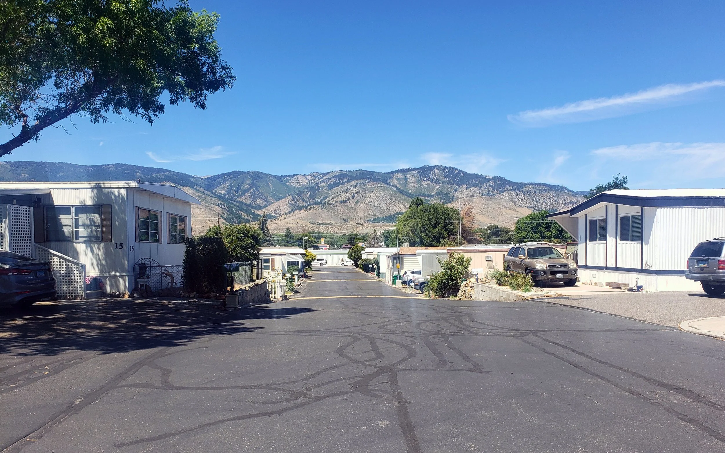 A mobile home park with rows of white trailer homes on either side of a paved road, with mountains in the background under a clear blue sky.