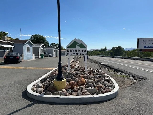 Sign for Rio Vista located at 4175 W. 4th Street, with stone decoration and a lamppost in the foreground, nearby small buildings and parked cars, along a stretch of road under clear blue sky.