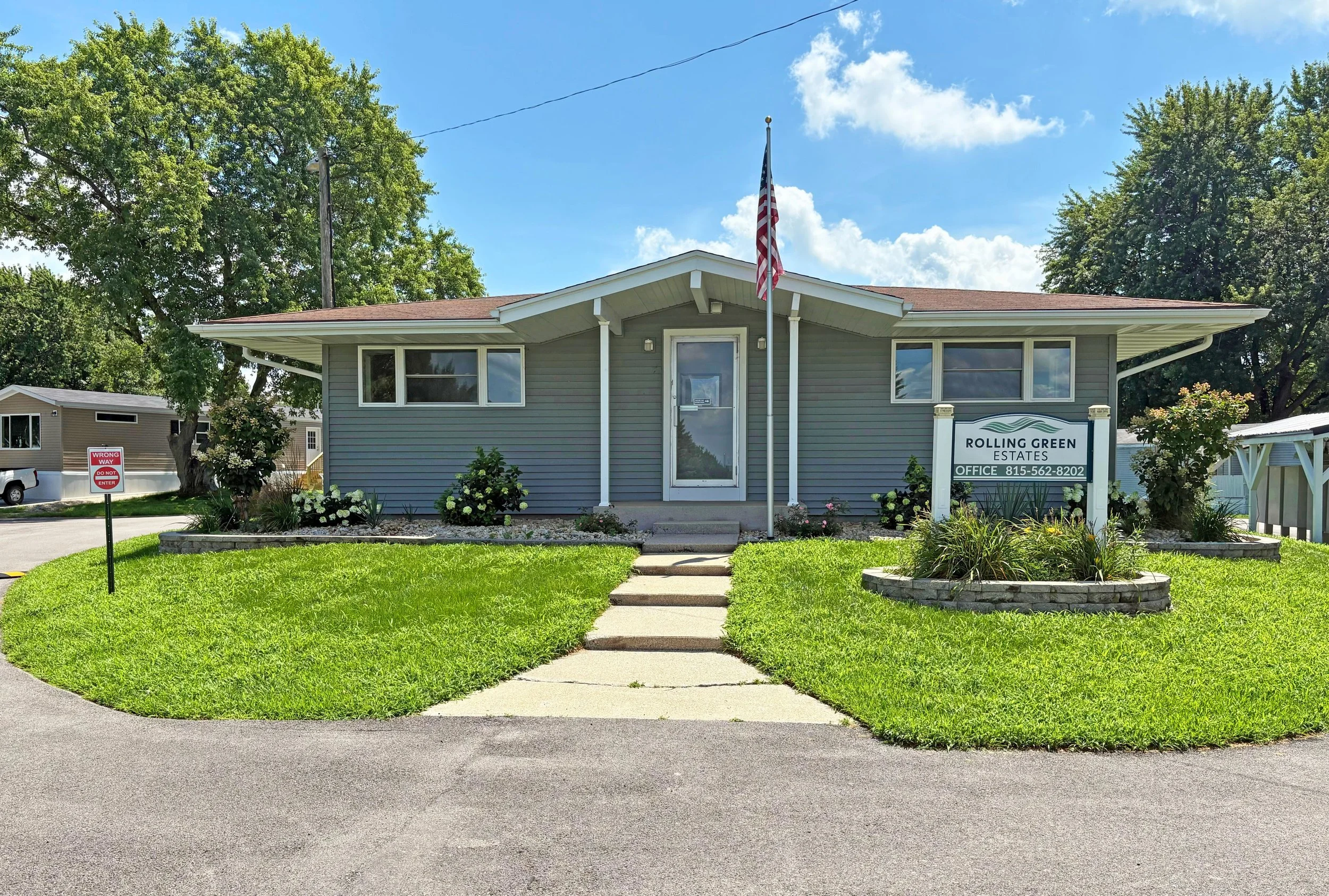 A single-story grey house with white trim, front steps, and an American flag outside. There is a sign for Rolling Green Estates and well-maintained lawn and landscaping.