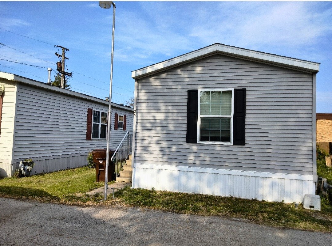 A small, gray, single-story house with black window shutters and white trim, featuring a window on the front wall, with a grassy yard and a sidewalk in front, a small set of stairs leading to the door, and a clear blue sky overhead.
