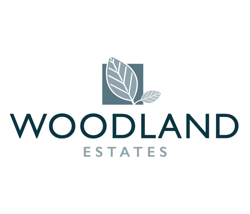 Logo for Woodland Estates featuring a stylized leaf inside a blue square. The word 'WOODLAND' is written below in large black letters, with 'ESTATES' in smaller gray letters underneath.