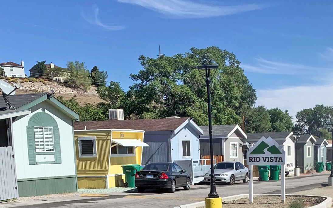 A street with small houses, a streetlamp, parked cars, and a sign that says Rio Vista, with green recycling bins visible.