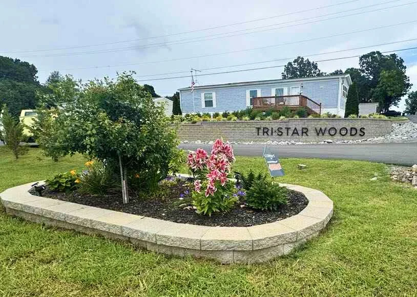 A landscaped garden bed with pink lilies and green shrubs, bordered by stone edging, in front of a sign that reads 'TRISTAR WOODS' with a house on a hill in the background.