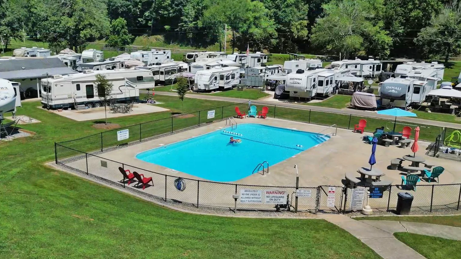 A swimming pool area surrounded by a fence with red and blue chairs and umbrellas, adjacent to a campground with numerous RVs and trailers, and a background of green trees.