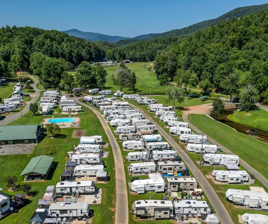 A large campground with numerous RV trailers parked in neat rows on grassy plots, surrounded by lush green trees and rolling hills under a clear blue sky.