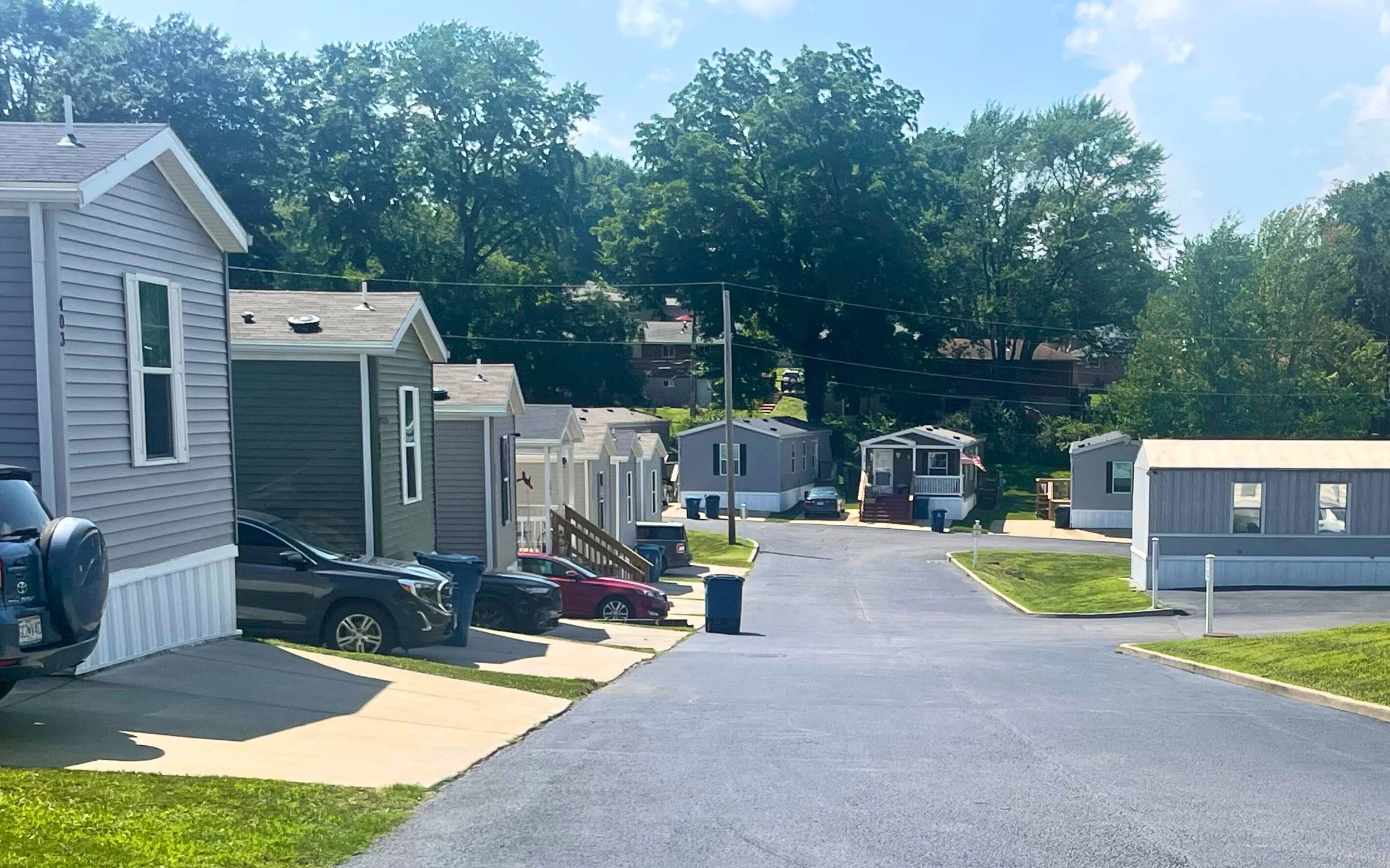 A mobile home park with several manufactured homes lined along driveways, some cars parked in front, green lawns, and large trees in the background under a bright blue sky.
