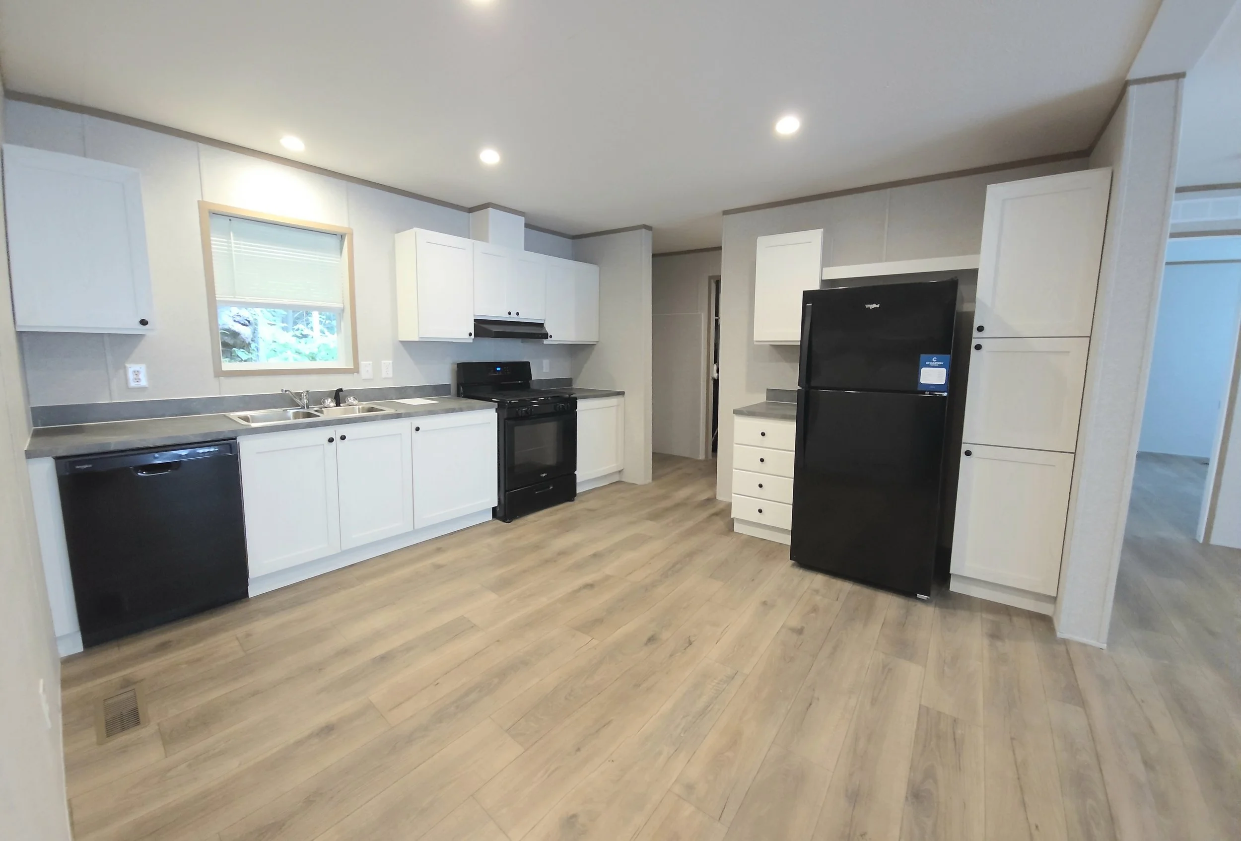 A modern kitchen with white cabinets, black appliances, and light wood flooring. It features a window above the sink, a black refrigerator, stove, dishwasher, and multiple drawers and cabinets for storage.