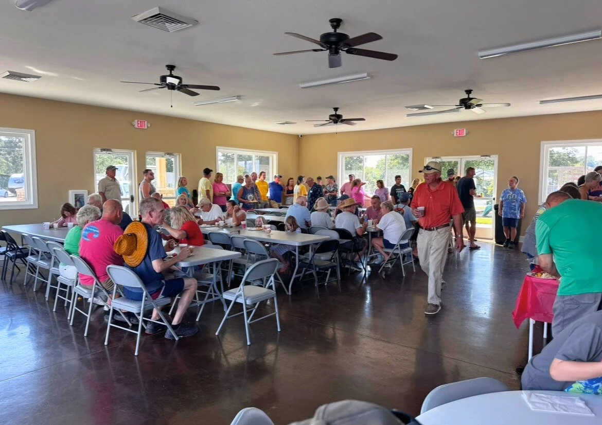 People gathered in a bright, spacious room with beige walls, sitting at long tables and standing in line, enjoying a meal or socializing during a community event or gathering.