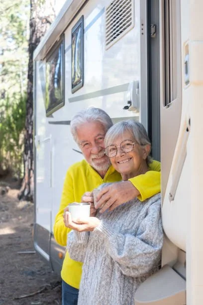 An elderly man and woman smiling and hugging outside a white camper trailer, with the man holding a mug.