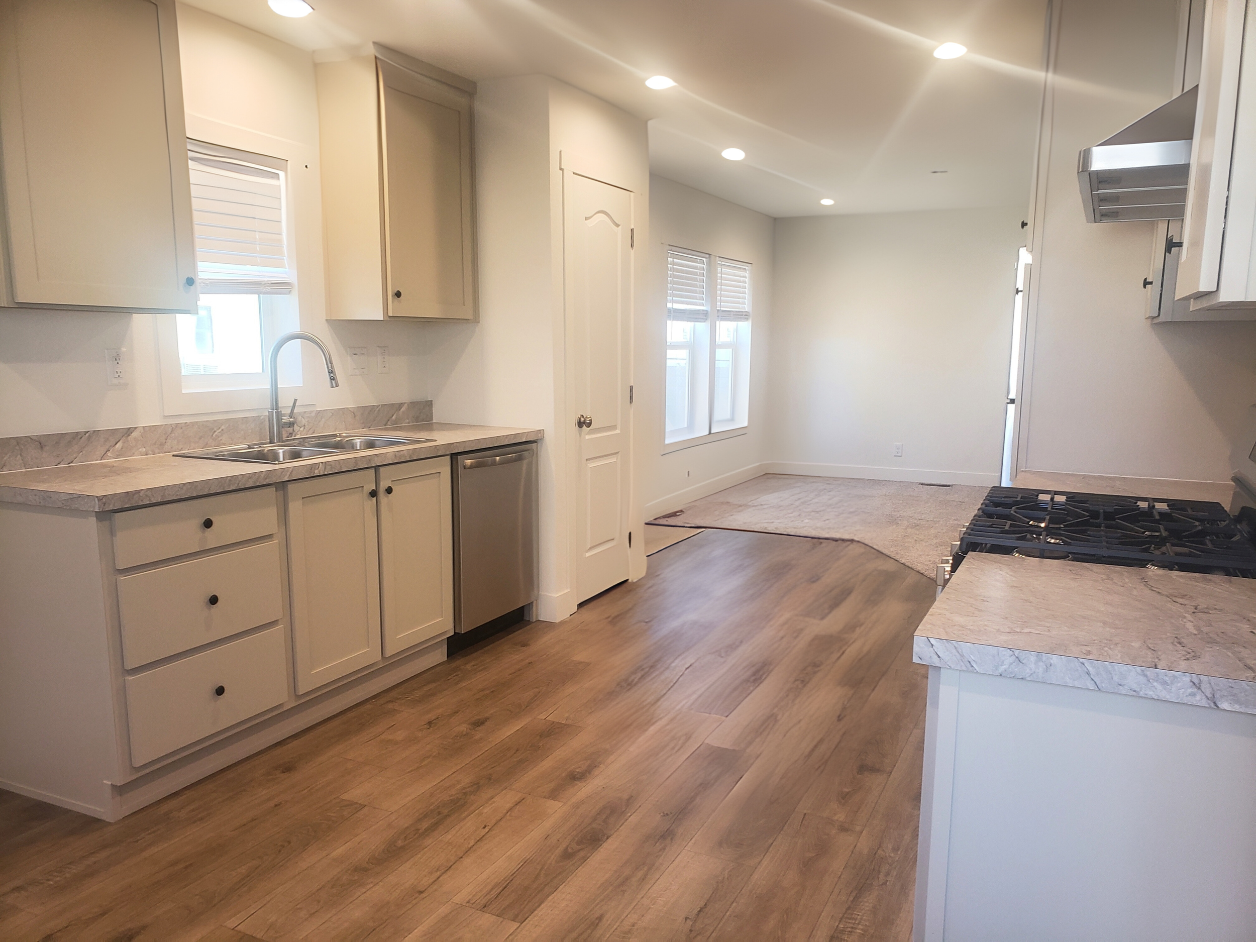 Kitchen with white cabinets, marble countertops, stainless steel sink, stove, and small window, hardwood floors, and a dining area with multiple windows and beige carpet.
