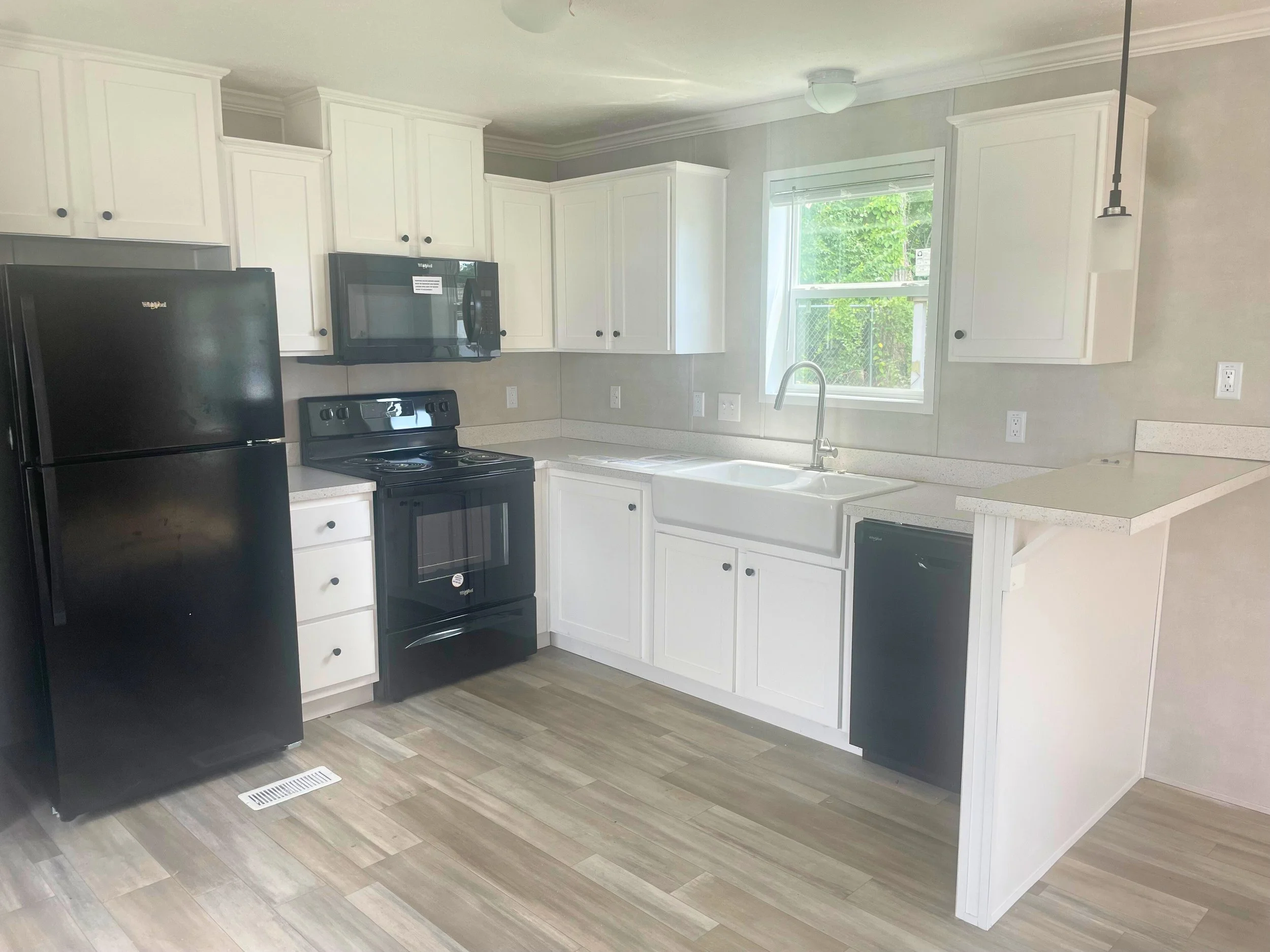 The kitchen has white cabinets, black appliances, a black refrigerator, stove, microwave, and dishwasher, with a window above the sink and light-colored wood flooring.