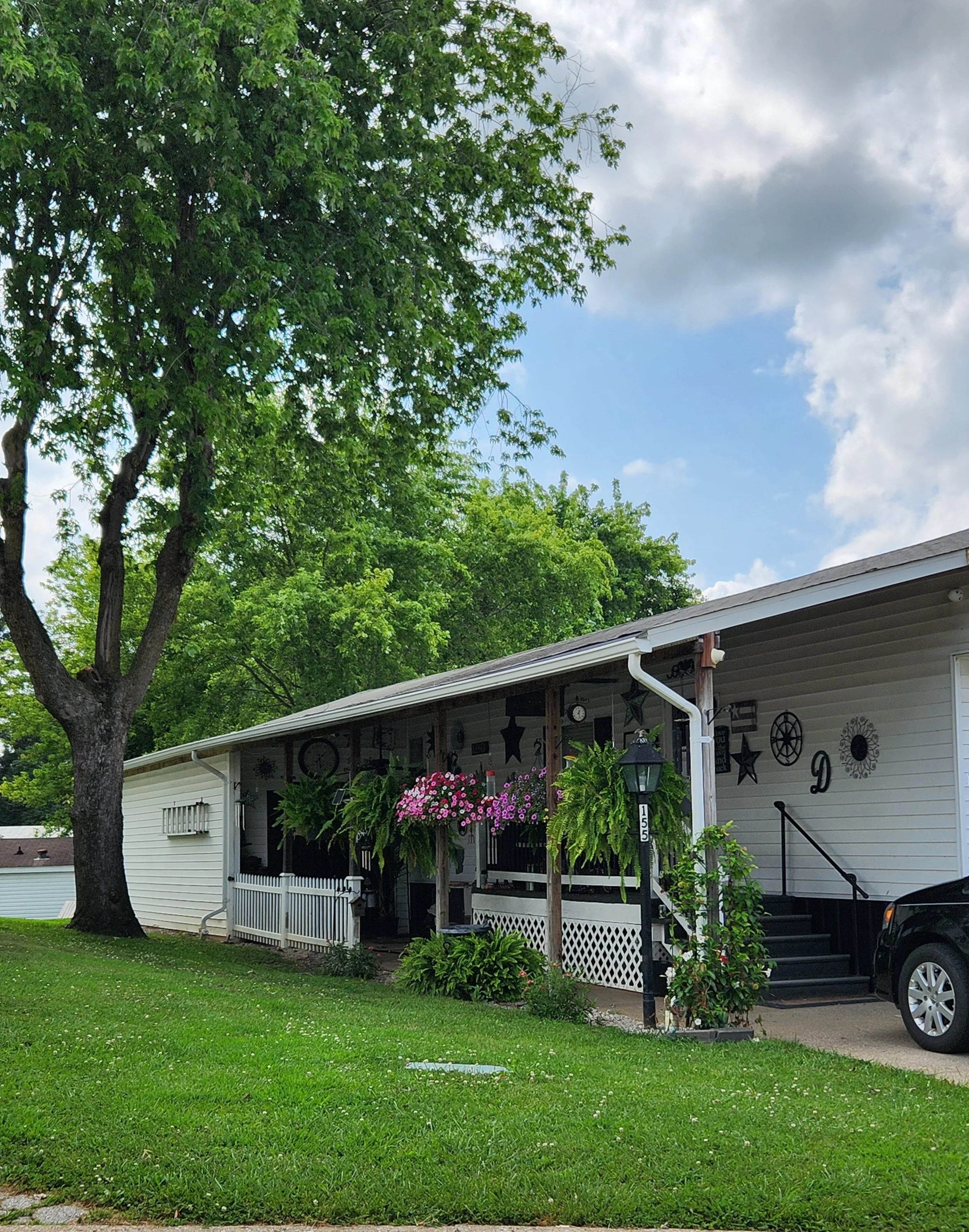 Front of a white house with a porch decorated with hanging plants and pink flowers, black stars, and outdoor wall art, with a large tree and green lawn in front, under a partly cloudy sky.