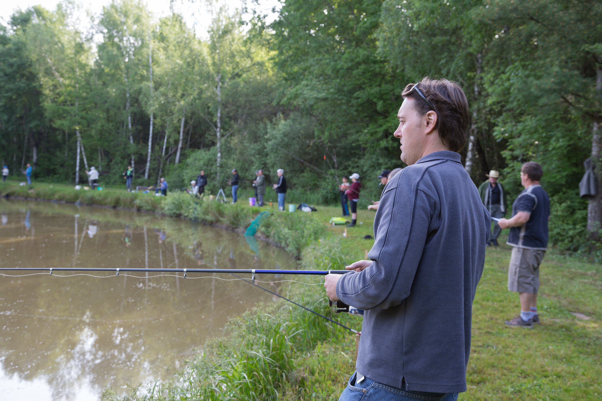 People fishing along the edge of a pond in a wooded area during daytime.