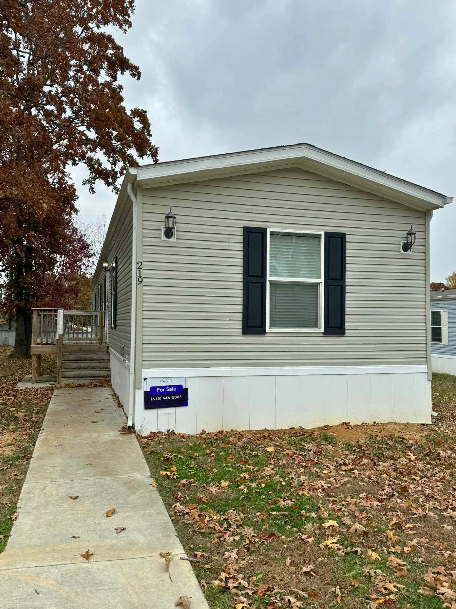Mobile home with beige siding and black shutters for sale, with a blue for sale sign on the front, and a porch with stairs on the left side, surrounded by fallen autumn leaves.