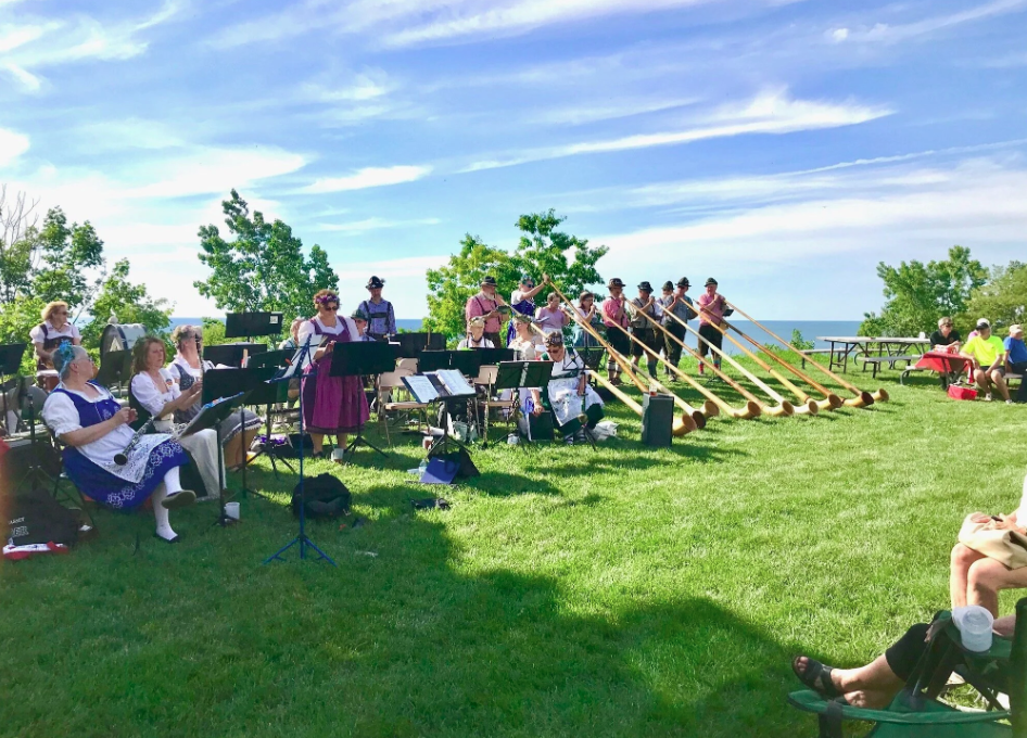 A group of musicians performing outdoors with horns and other instruments, wearing traditional clothing, under a sunny sky with a view of the ocean in the background.