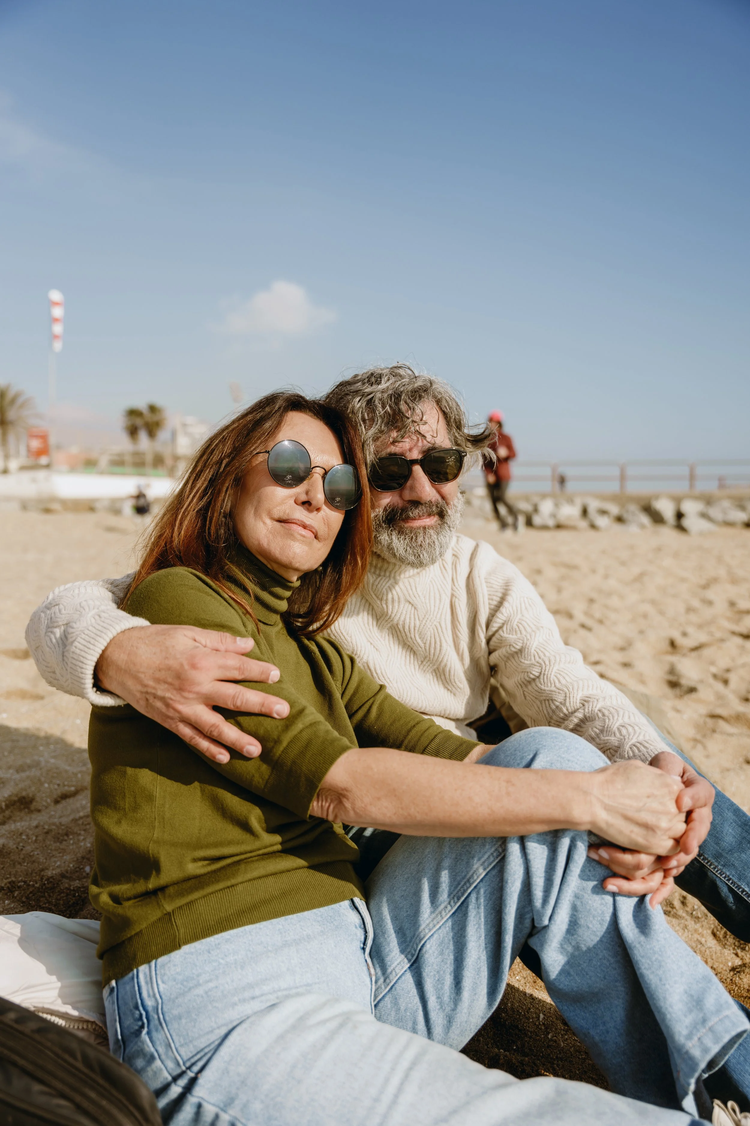 A middle-aged man and woman sitting on the sand at a beach, embracing and wearing sunglasses. The woman has brown hair and a green top, while the man has gray hair, a beard, and a cream sweater. In the background, there are people walking, a promenad