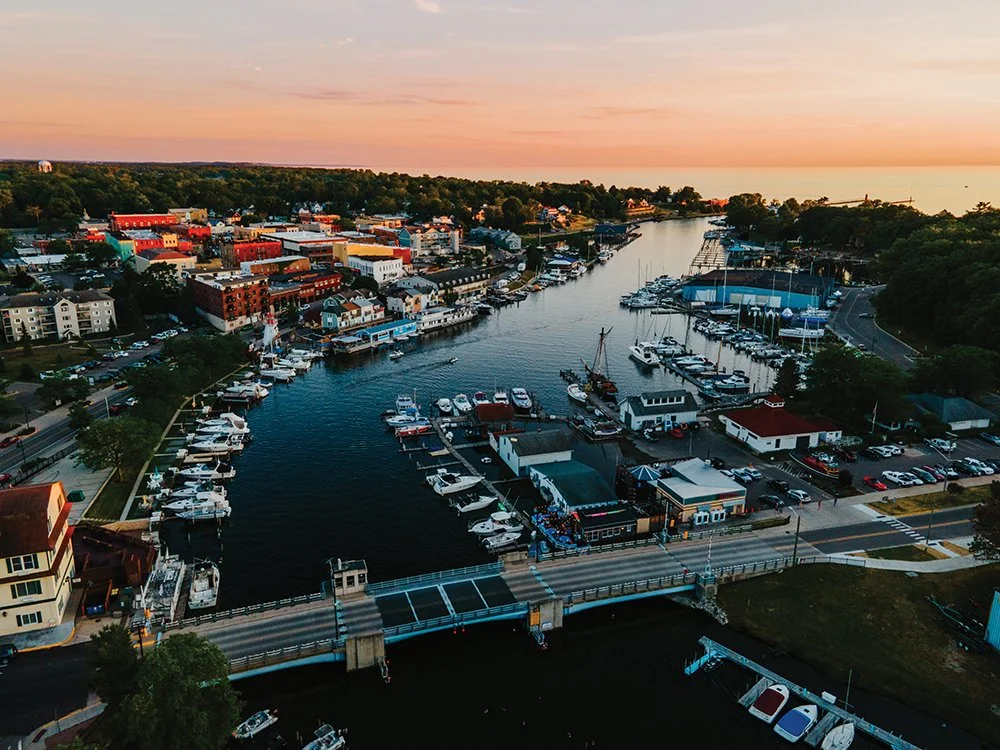 Aerial view of a marina with boats docked along a waterway, surrounded by colorful buildings and a bridge, at sunset.