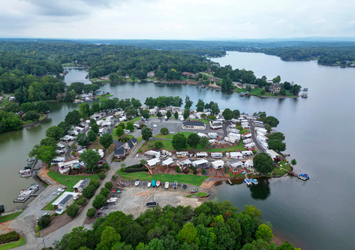 Aerial view of a lakeside residential community with houses, boats, and docks surrounded by green trees and water, under an overcast sky.