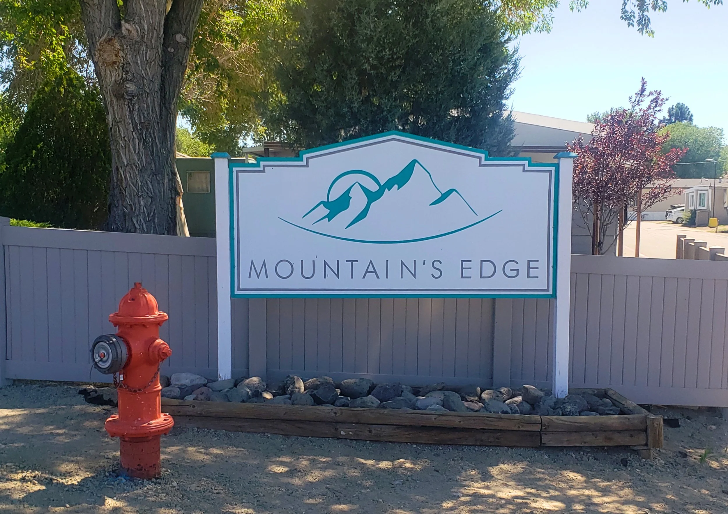 A sign reading 'Mountain's Edge' with a mountain graphic, placed in front of trees and houses, with a red fire hydrant nearby.