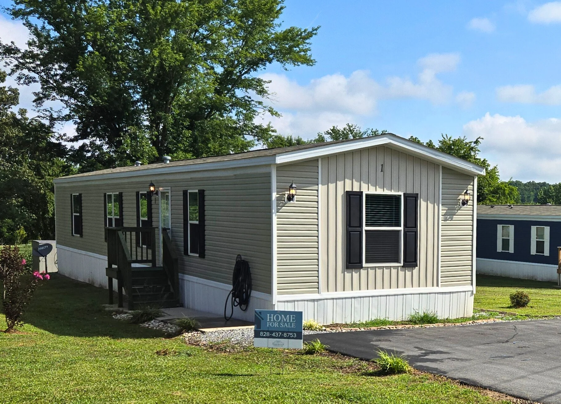 A beige manufactured home with black shutters, a small wooden porch, and a yard, for sale as indicated by a sign in the front yard.