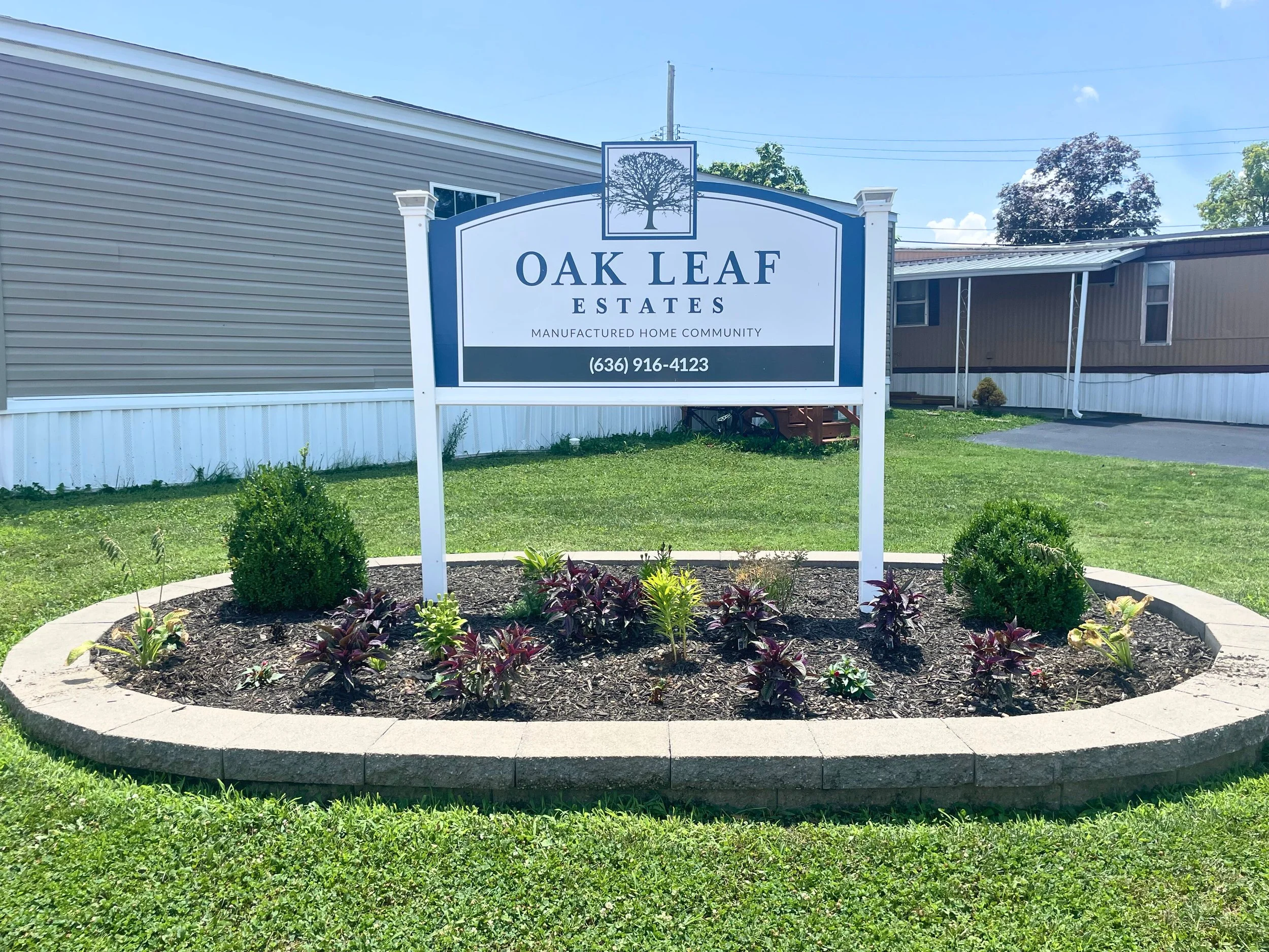 Sign for Oak Leaf Estates manufactured home community in a landscaped yard with bushes and flowers, next to mobile homes.