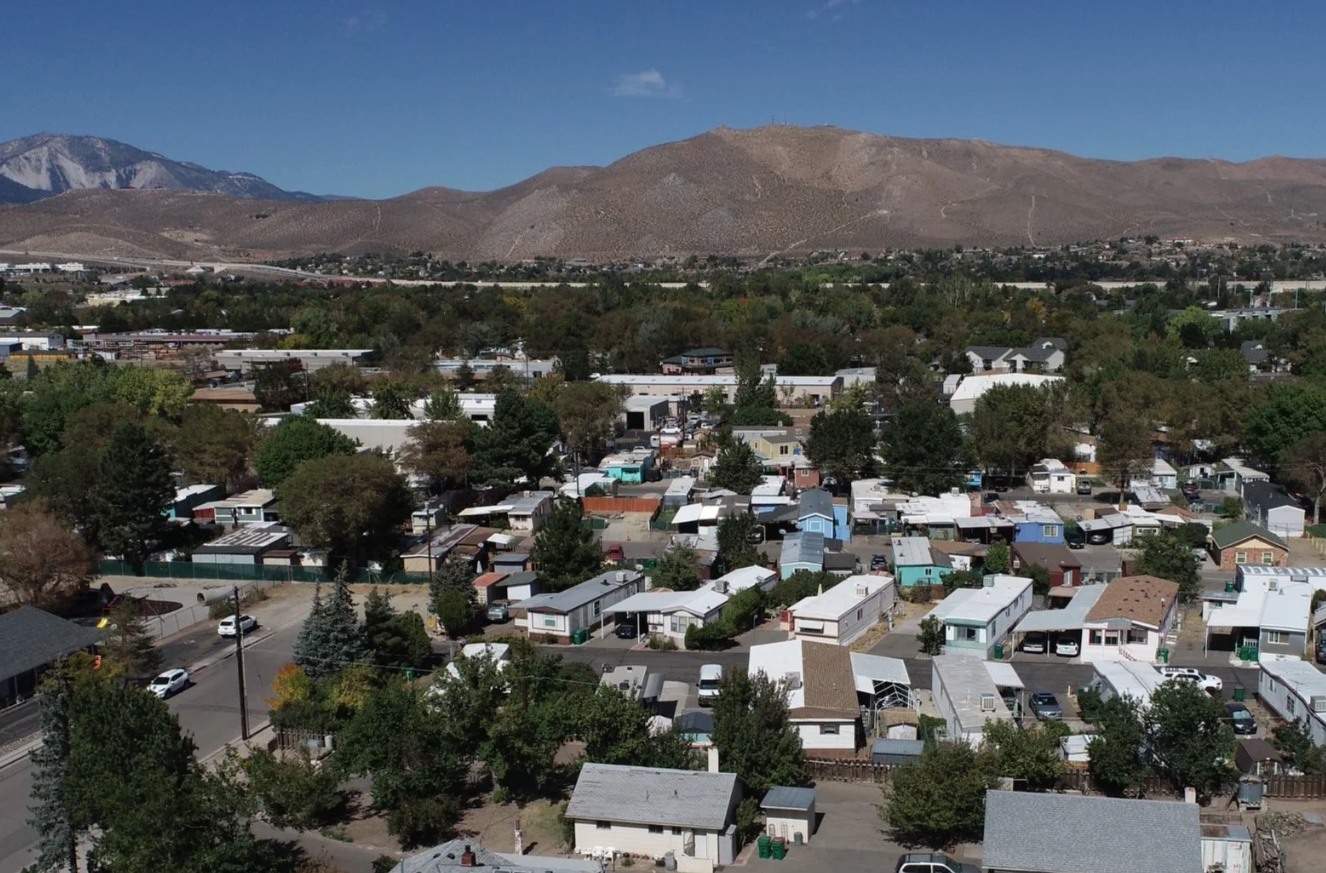 Aerial view of a suburban neighborhood with small houses, trees, and streets, set against a backdrop of mountains under a blue sky.