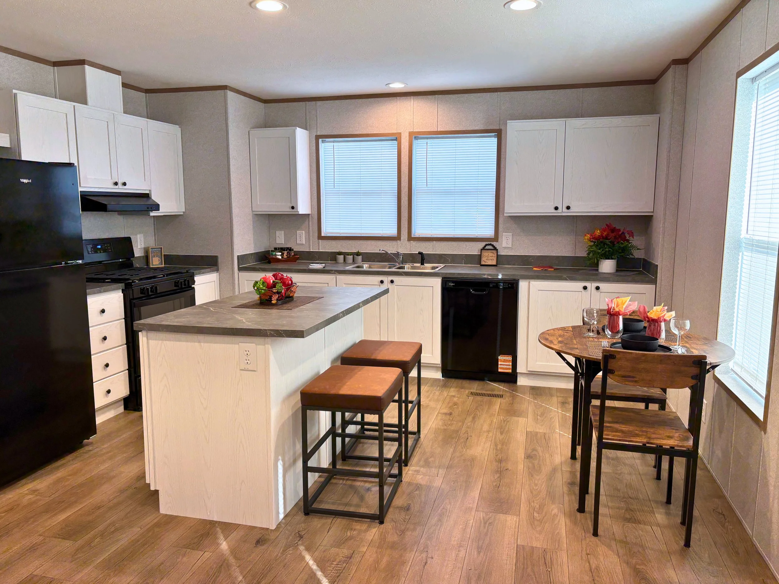 Kitchen with white cabinets, black appliances, a small island with two stools, a dining table set for three, wood flooring, and two windows above the sink.