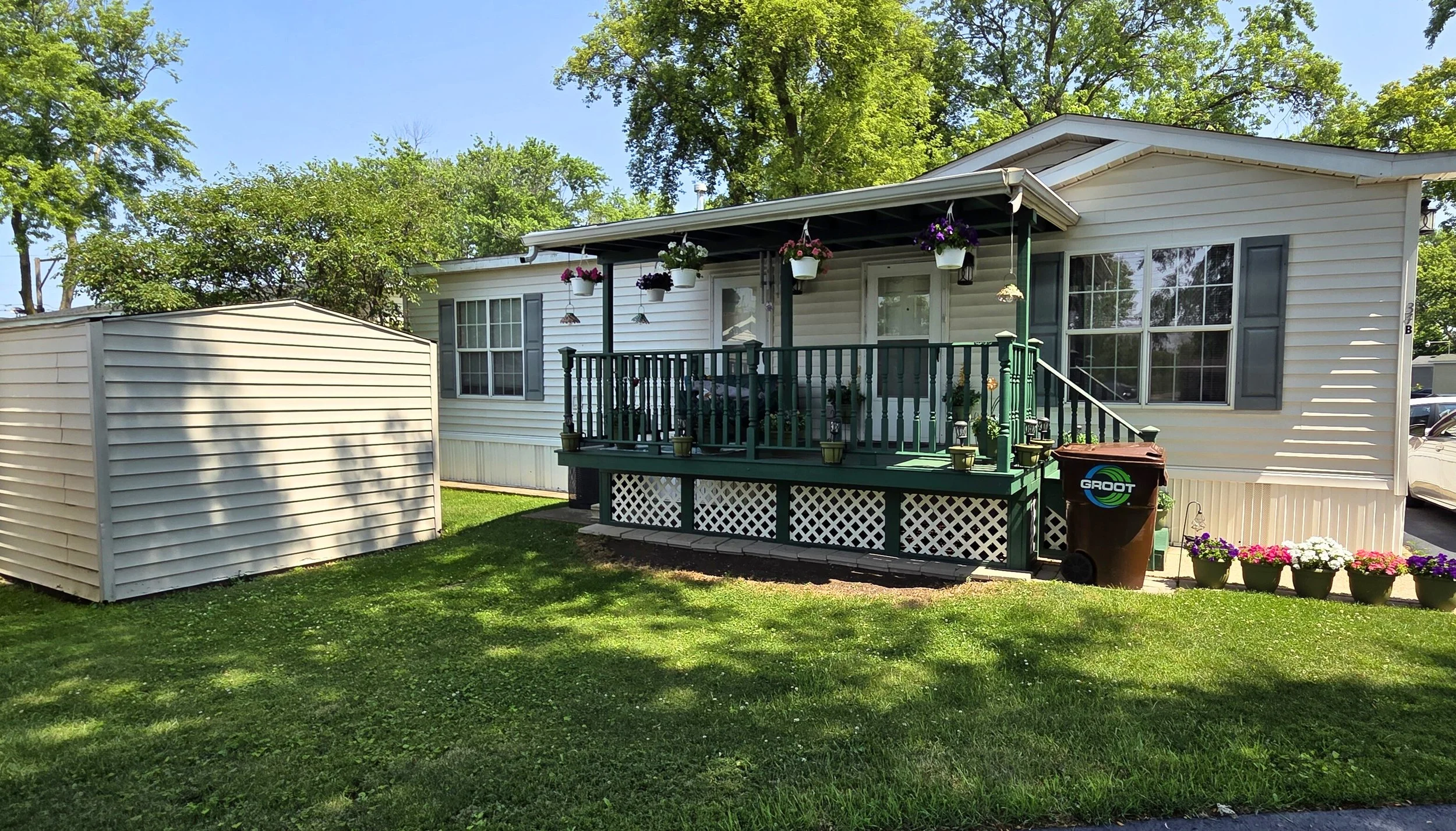 A small house with a front porch decorated with hanging flower baskets and potted plants, a shed on the left, and a green lawn with trees in the background.