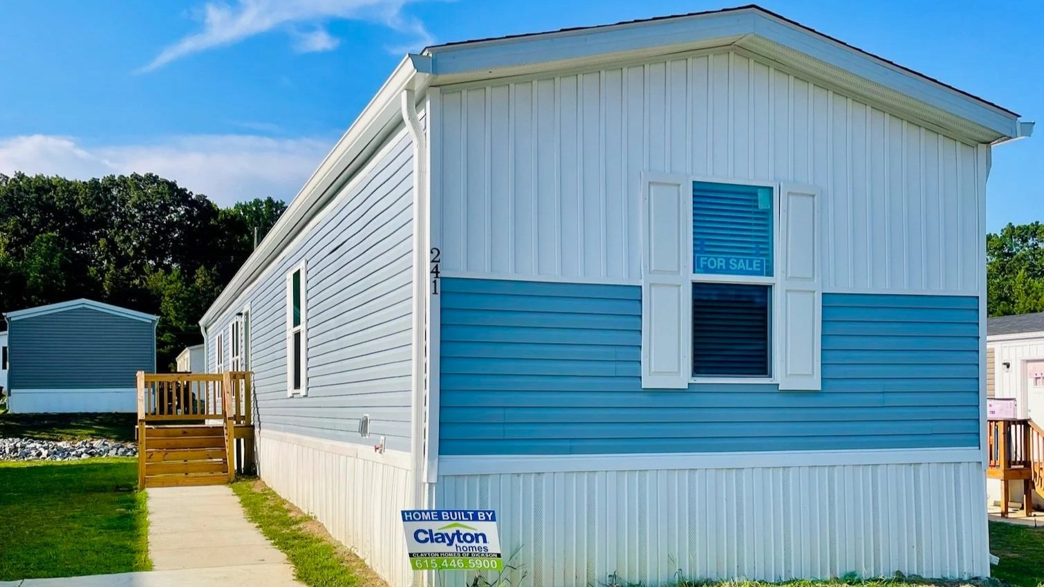 Blue and white manufactured home with a 'For Sale' sign in the window, a small porch with steps on the side, and a sign indicating it was built by Clayton Homes.