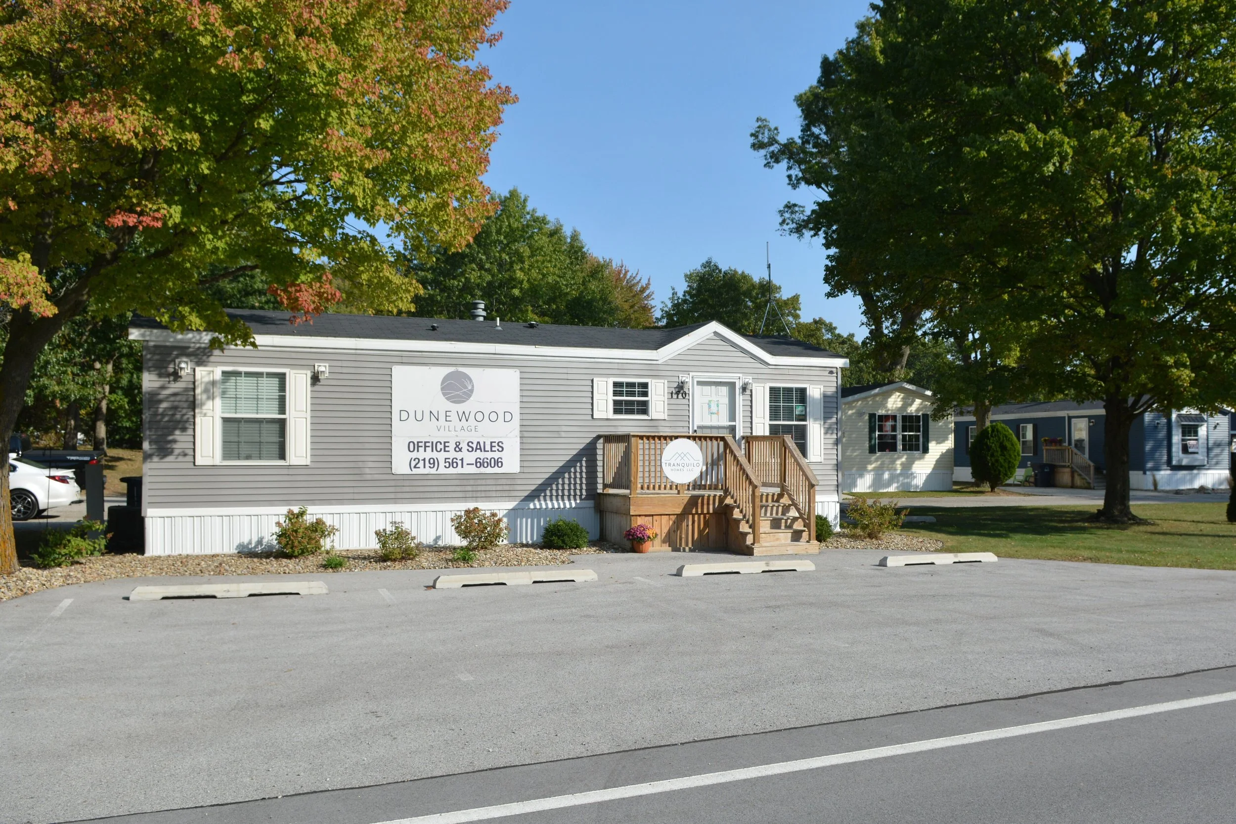 Single-story gray building with white trim and a small wooden porch, surrounded by trees with green and some autumn-colored leaves, in a parking lot with marked spaces.