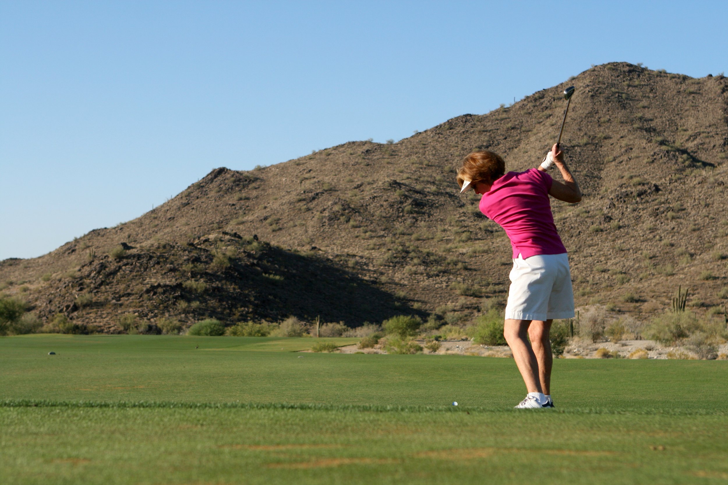 Senior woman in pink shirt and white shorts playing golf on a course with desert mountains in the background.