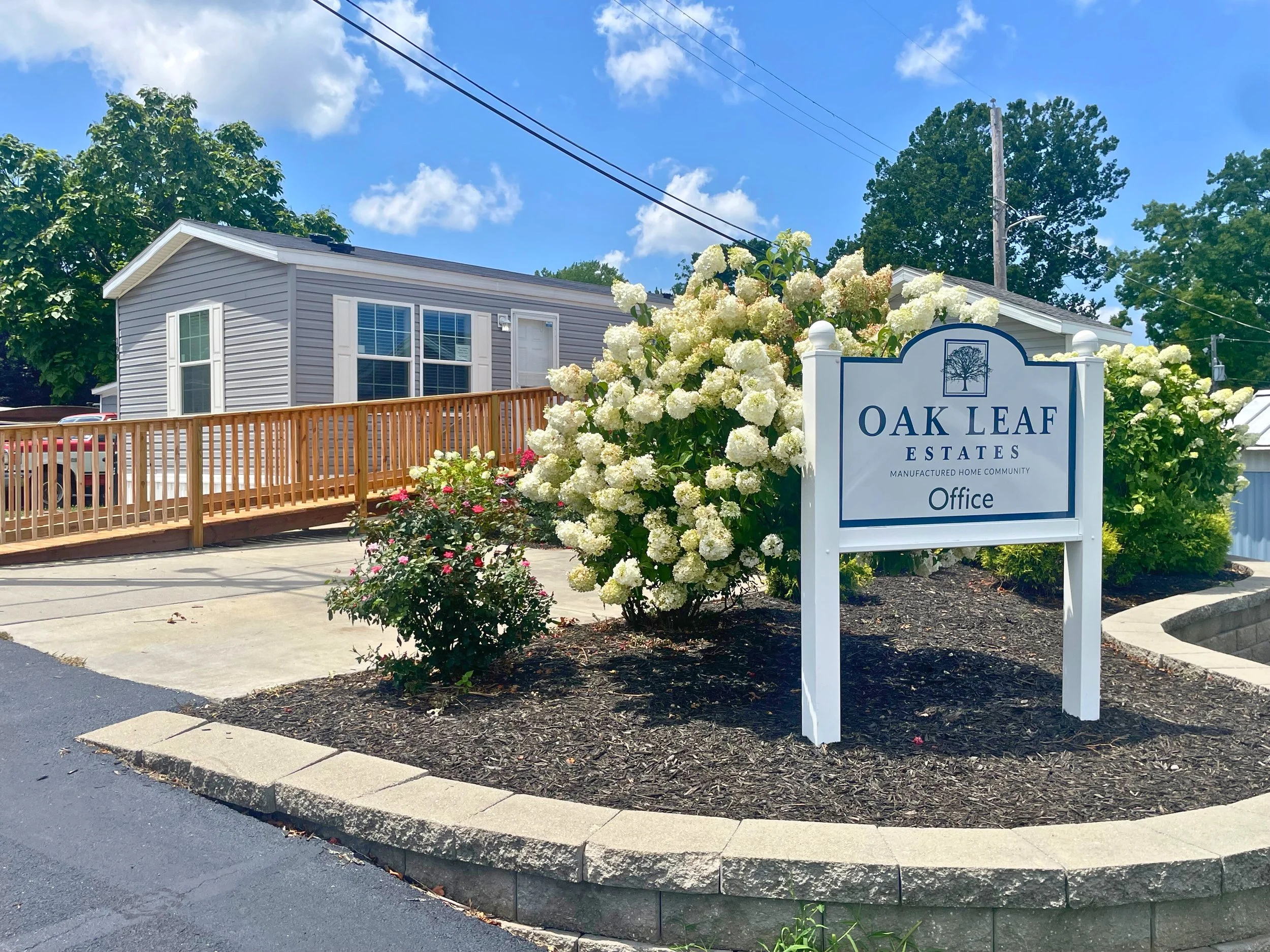 Sign for Oak Leaf Estates community office surrounded by white hydrangea flowers and other shrubs with a mobile home and driveway in the background under a blue sky with clouds.