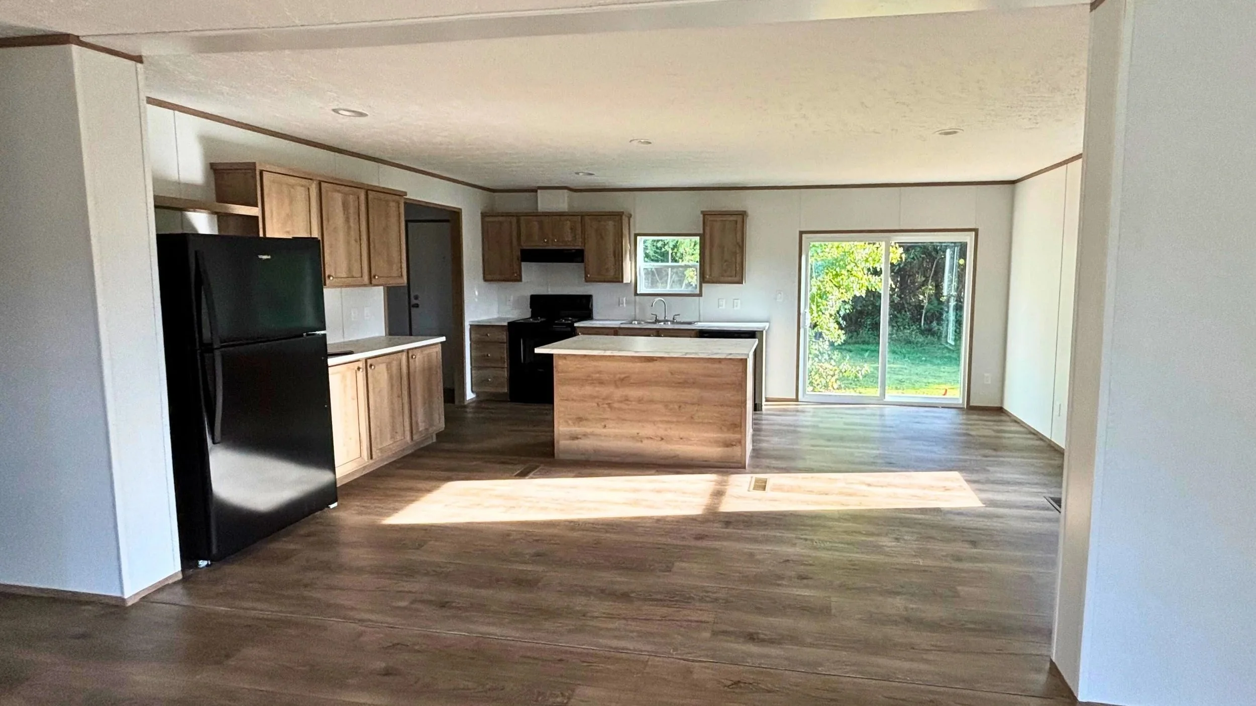 Empty kitchen with black refrigerator, wooden cabinets, island with countertop, black stove, and sliding glass door leading to a yard.
