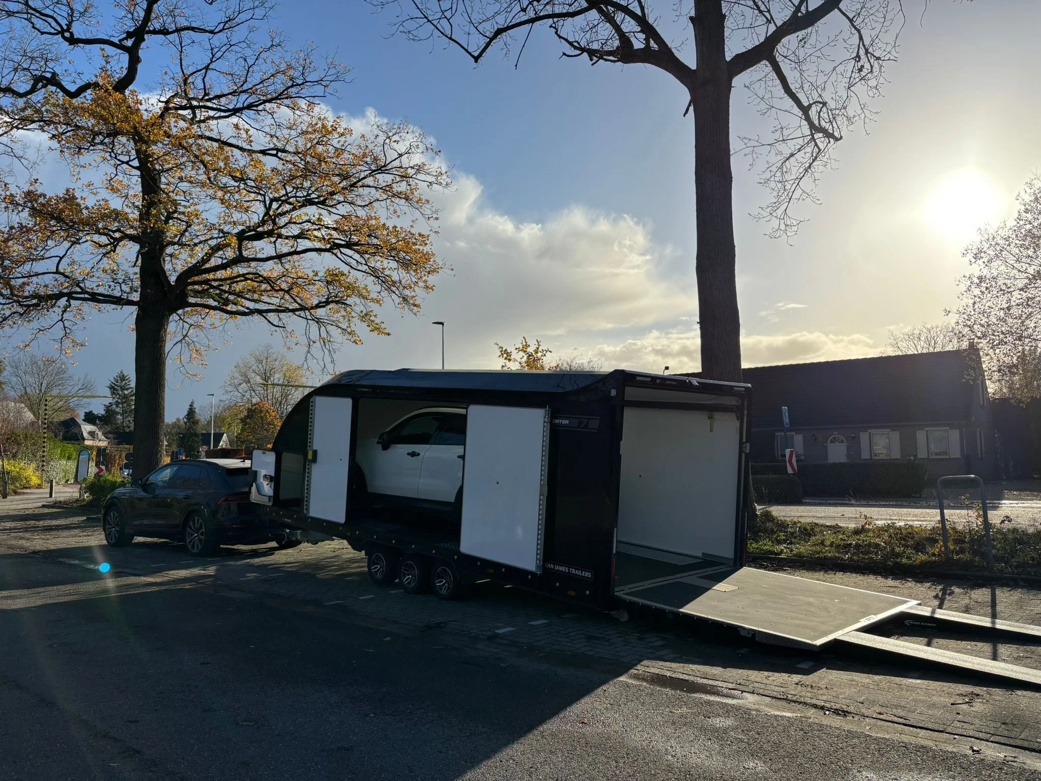 A Brian James RT7 car transport trailer with a white SUV car inside, parked on a street.