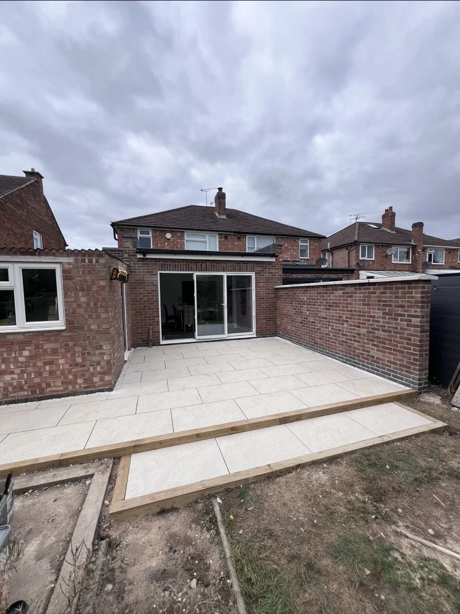 Backyard patio with large light-colored tiles, partially bordered by wooden framing, attached to a brick house with sliding glass doors, neighboring brick houses under cloudy sky.