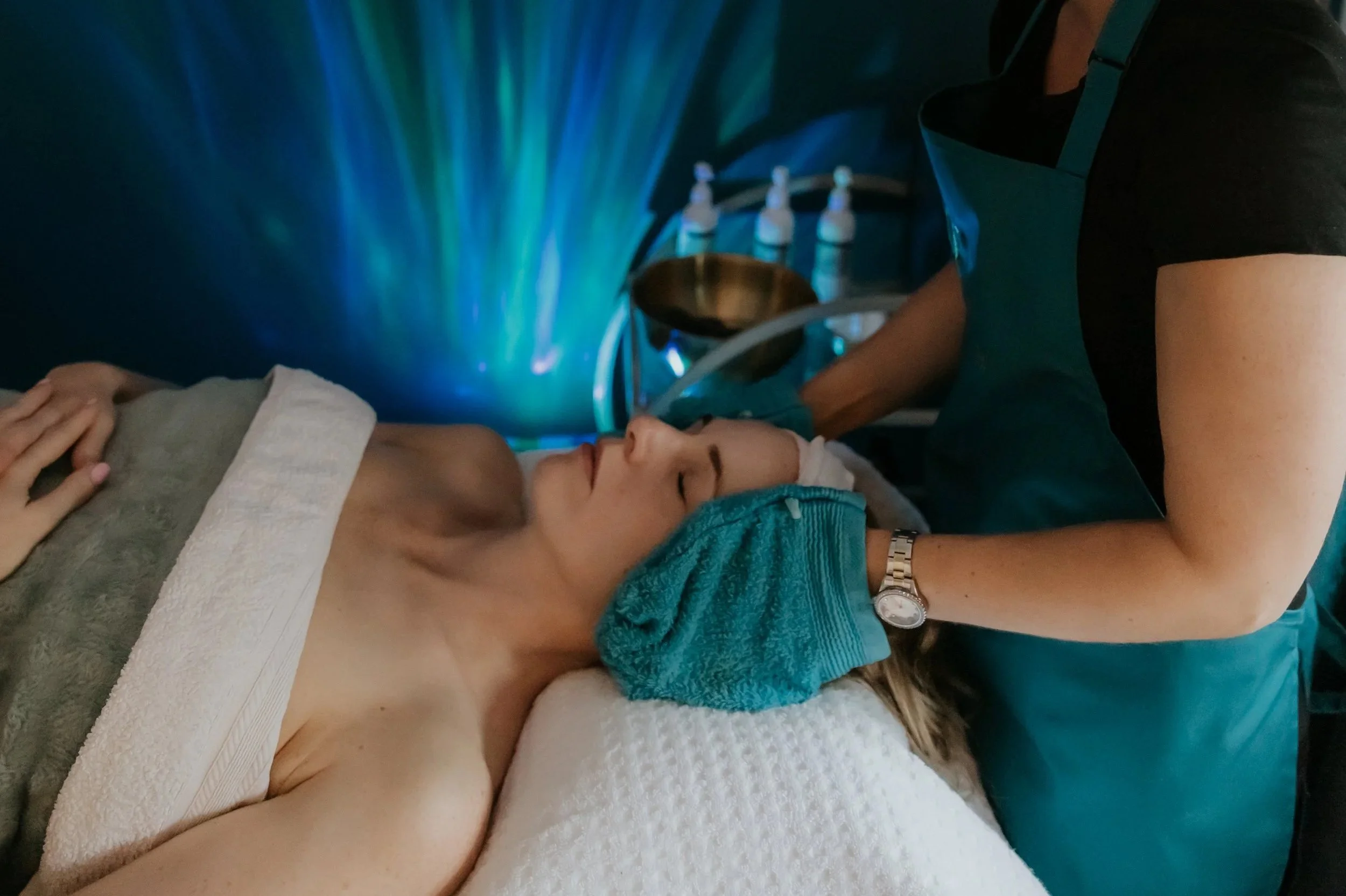 Woman laying on a treatment bed. Therapist standing above holding blue face towels to the lady's face