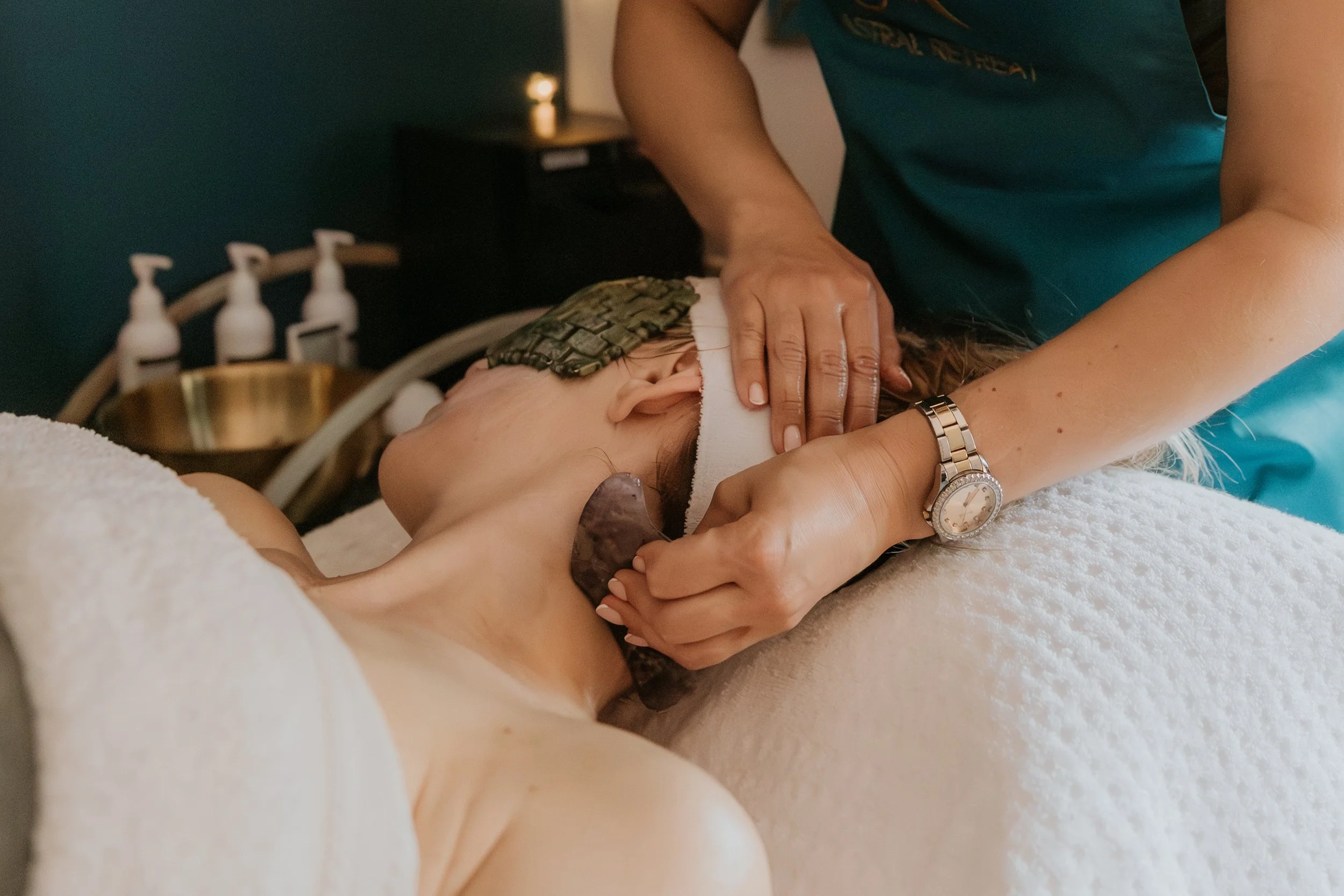 Spa therapist performing a gua sha facial treatment on a client lying under a towel with skincare bottles nearby