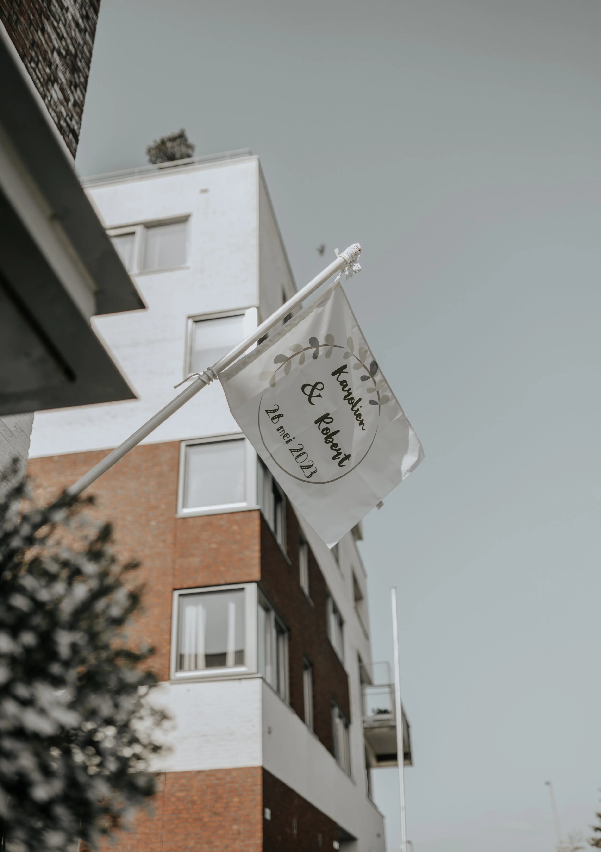 Een vlag met de tekst 'Karolien & Robert' en de datum 26 mei 2023 hangt uit een gebouw met een moderne, gemengde witte en roodbruine gevel, onder een grijze lucht.