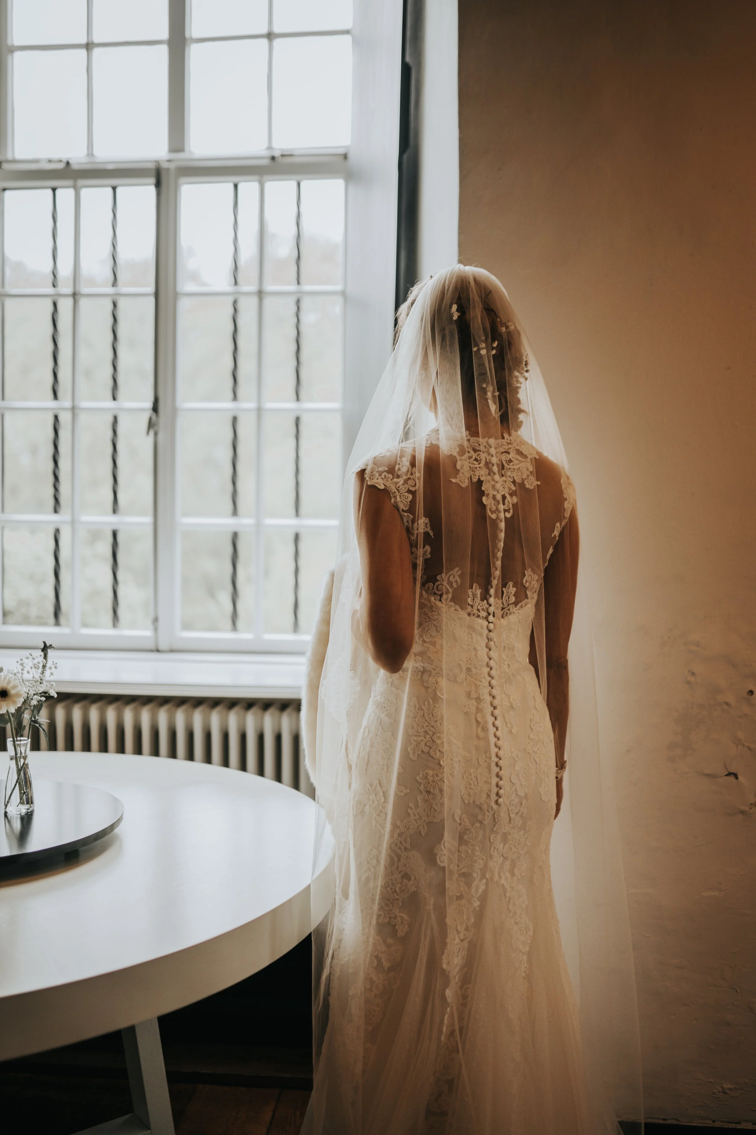 Bruidsvrouw in witte trouwjurk en sluier staat binnen naast een groot raam met matglas, met een ronde tafel met bloemen voor haar.